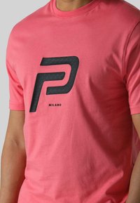 Ružičasta pamučna majica s kratkim rukavima koja ima veliki crni teksturirani logo "P" sa riječi "MILANO" ispod. Okrugli izrez.