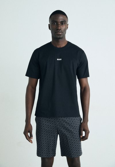 T-shirt noire en coton avec un logo sur la poitrine, associée à un short gris présentant un motif géométrique. Le modèle se tient devant un fond sobre.
