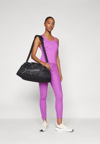 Svart duffelbag med hvite detaljer, båret på skulderen. Modell i lilla tank topp og mønstrete tights, står oppreist med nøytral bakgrunn.