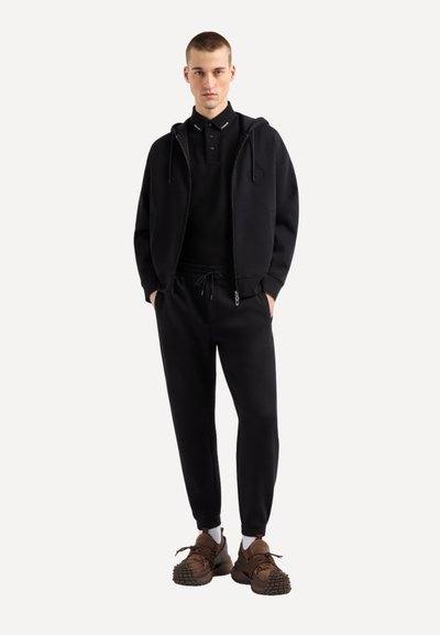 Sweat à capuche noir zippé avec cordon de serrage, porté par-dessus une chemise polo noire. Un jogging noir resserré et des baskets marron complètent le look.
