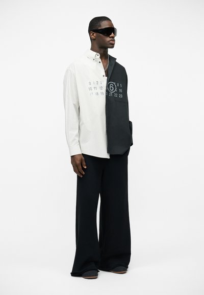 Άνδρας που φοράει υπερμεγέθη ασπρόμαυρο πουκάμισο με κουμπιά και τυπωμένους αριθμούς, φαρδιά μαύρη παντελόνα και μεγάλα μαύρα γυαλιά ηλίου.