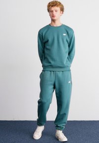 Teal sweatshirt en bijpassende broek, beide gemaakt van zachte stof. De sweatshirt heeft een ronde halslijn en logo. De broek heeft een comfortabele pasvorm en een elastische tailleband.