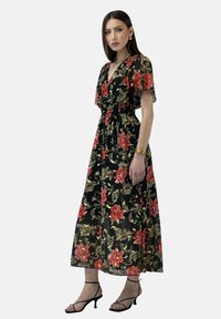 Vestido largo negro con estampado floral en rojo y verde, con mangas cortas con vuelo, cintura ceñida y una silueta fluida. Combinado con tacones negros con tiras.