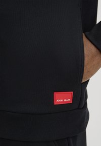 Zwarte geribbelde sweatshirts met een rood label met de tekst "JOOP! JEANS" aan de zijkant, uitgevoerd met een gestructureerd oppervlak en aansluitende manchetten.