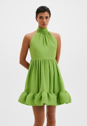 Femme portant une robe sans manches vert citron à col halter, taille froncée et ourlet volanté, debout devant un fond blanc.