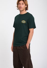 Jeune homme aux cheveux blonds mi-longs portant un t-shirt vert foncé avec un logo soleil jaune et un pantalon ample marron foncé, les mains dans les poches.