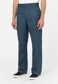 Pantalons taille haute, jambes droites en bleu sarcelle, fabriqués en tissu lisse avec des plis avant. Associés à des chaussures noires à enfiler.