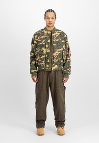 Camouflage bomber jacket in green and brown, ribbed cuffs, zip closure. Olive cargo pants with side pockets, worn with beige boots.
