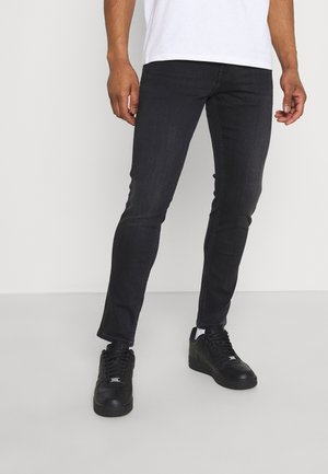 Hombre con vaqueros ajustados negros, zapatillas negras y camisa blanca de pie frente a un fondo blanco.