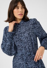 Robe à boutons en bleu marine foncé avec un motif abstrait bleu clair. Présente des manches bouffantes et une ceinture en tissu à la taille pour un look ajusté.