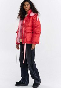Czerwona kurtka puchowa z różowymi akcentami, zapinana na zamek błyskawiczny, z ściągaczami i bocznymi kieszeniami, noszona na kolorowym swetrze i ciemnych dżinsach.