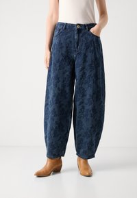 Laiad denim püksid tumeda sinise lillemustriga, millel on nupplukustus, vööniidid ja küljetaskud. Kannab pruuni pahkluu saapaga.