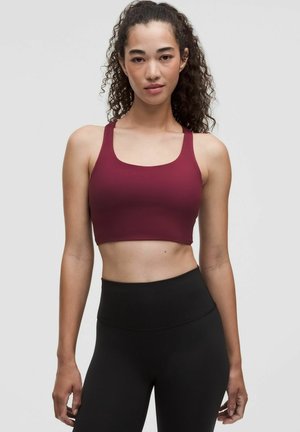 Giovane donna con capelli ricci indossa un reggiseno sportivo bordeaux e leggings neri a vita alta, in piedi davanti a uno sfondo semplice.