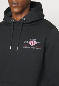 Homme portant un sweat à capuche noir avec le logo "GANT", un emblème de bouclier et le texte "American Sportswear" sur la poitrine, montrant le haut du torse et le cou.