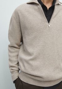 Maglione beige a coste con collo con zip, caratterizzato da una texture morbida e una silhouette aderente, indossato sopra una camicia scura a quadri.
