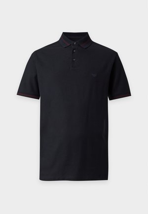 Junger Mann steht, trägt ein schwarzes kurzärmeliges Polohemd, hellblaue Jeans mit weitem Bein und braune Freizeitschuhe vor weißem Hintergrund.