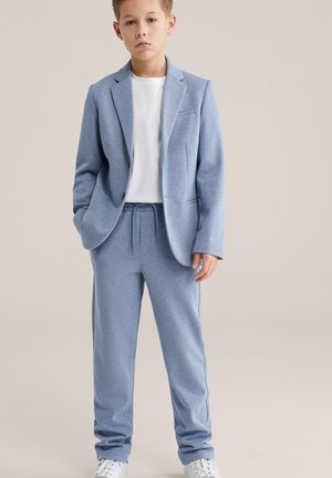 Világoskék öltönykabát és hozzáillő, gumis derékpánttal ellátott melegítőnadrág fehér póló fölött viselve, sima anyagból és szabott dizájnnal.