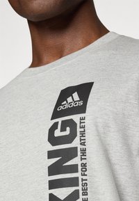 Grå bomullst-skjorte med en stor svart "KING" grafikk og "adidas" logo. Enkel design med rund hals og en myk, jevn tekstur.
