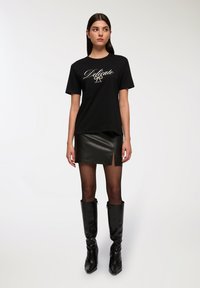 Czarna bawełniana koszulka z metalicznym napisem "Delikatny" i wzorem kokardy, połączona z czarną skórzaną mini spódnicą i wysokimi czarnymi botkami.