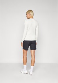 Pulover alb din tricot cu model de diamant, cu manșete cu nervuri, asortat cu pantaloni scurți negri. Modelul poartă adidași albi și șosete până la gleznă. Fundal simplu.