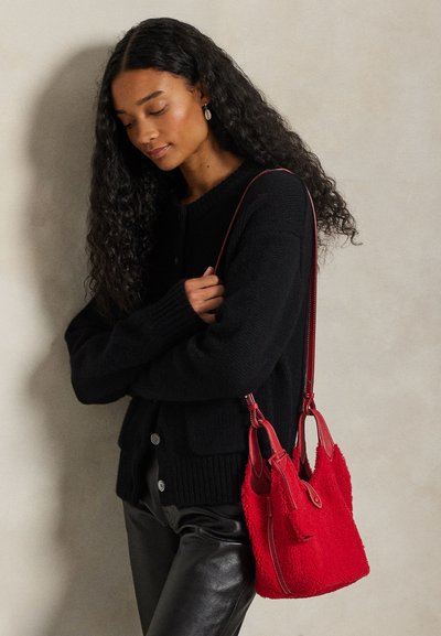 Červená kabelka z umělé kožešiny s koženými doplňky, kulatým tvarem a ramenním popruhem, doplněná o černý pletený kardigan a kožené kalhoty.