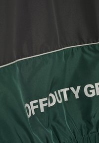 Dunkelgrüner und schwarzer Stoff mit weißer Paspelierung und teilweise sichtbarem, fettem weißen Text mit der Aufschrift "OFF DUTY GR".