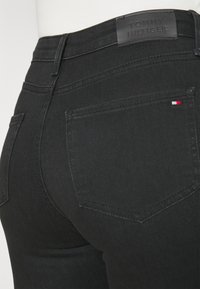 Primer plano de los bolsillos traseros de unos jeans negros de Tommy Hilfiger, mostrando la costura, el pequeño logo de la bandera en el bolsillo derecho y la etiqueta de cuero con la marca en la cintura.
