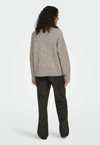 Femme avec des cheveux bouclés portant un pull en peluche gris et un pantalon noir large, debout et de dos sur un fond blanc uni.
