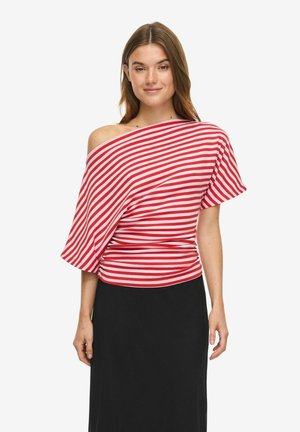 Kvinde iført rød og hvid diagonalt striped off-shoulder top og sort nederdel, stående mod en ensfarvet hvid baggrund.