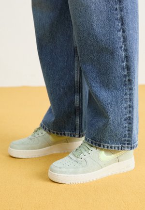 Persona con zapatillas de ante verde claro con suelas blancas y jeans azules, de pie sobre un suelo amarillo.