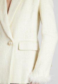 Blazer de lana blanca con cuello chal, cierre de botón de perla, un bolsillo lateral y acentos de pluma en los puños.