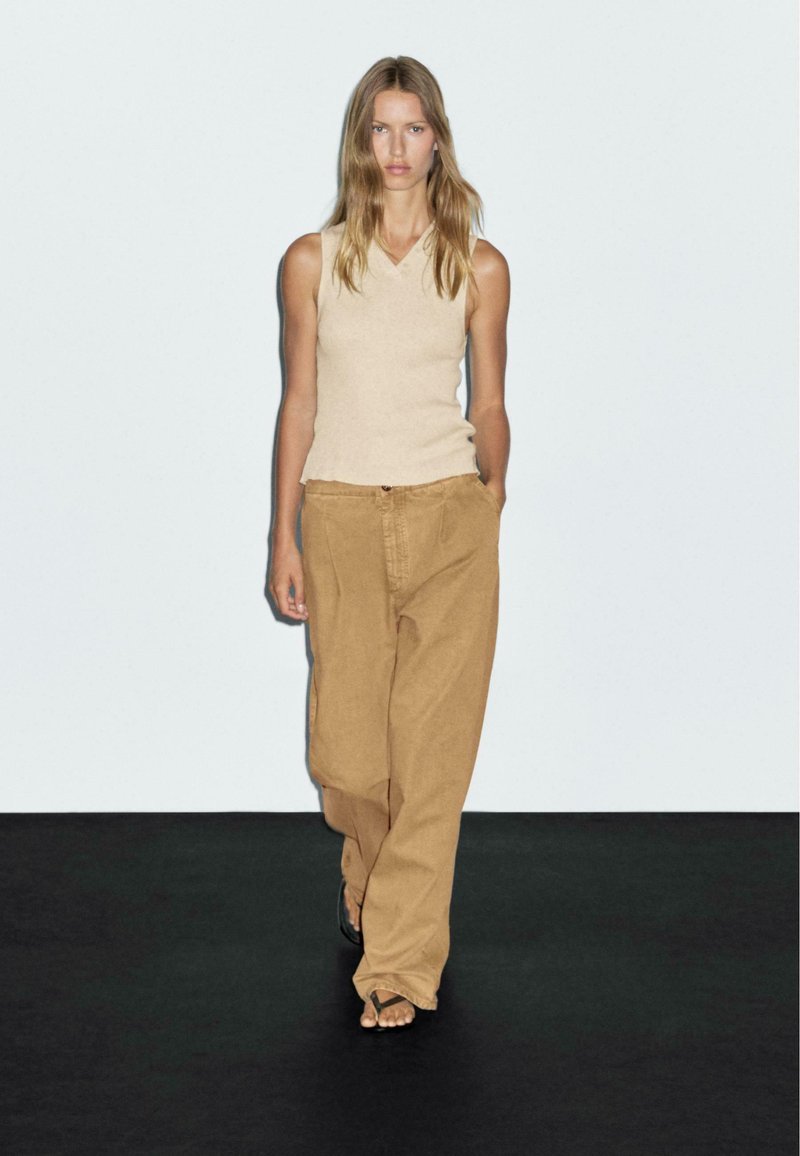 Woman walking in beige sleeveless top, loose tan pants, and black sandals against a plain white background and black floor.