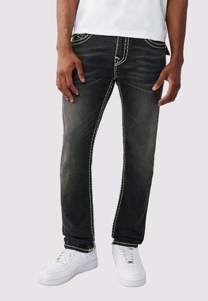 Mann trägt ausgebleichte schwarze Jeans mit weißer Kontrastnaht und weißen Turnschuhen, steht vor einem schlichten Hintergrund.