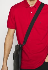 Punainen polo-paita, jossa on kaulus ja kolme nappia, ja pienellä logolla. Yhdistetty mustaan olkalaukkuun, jossa on sileä pinta.