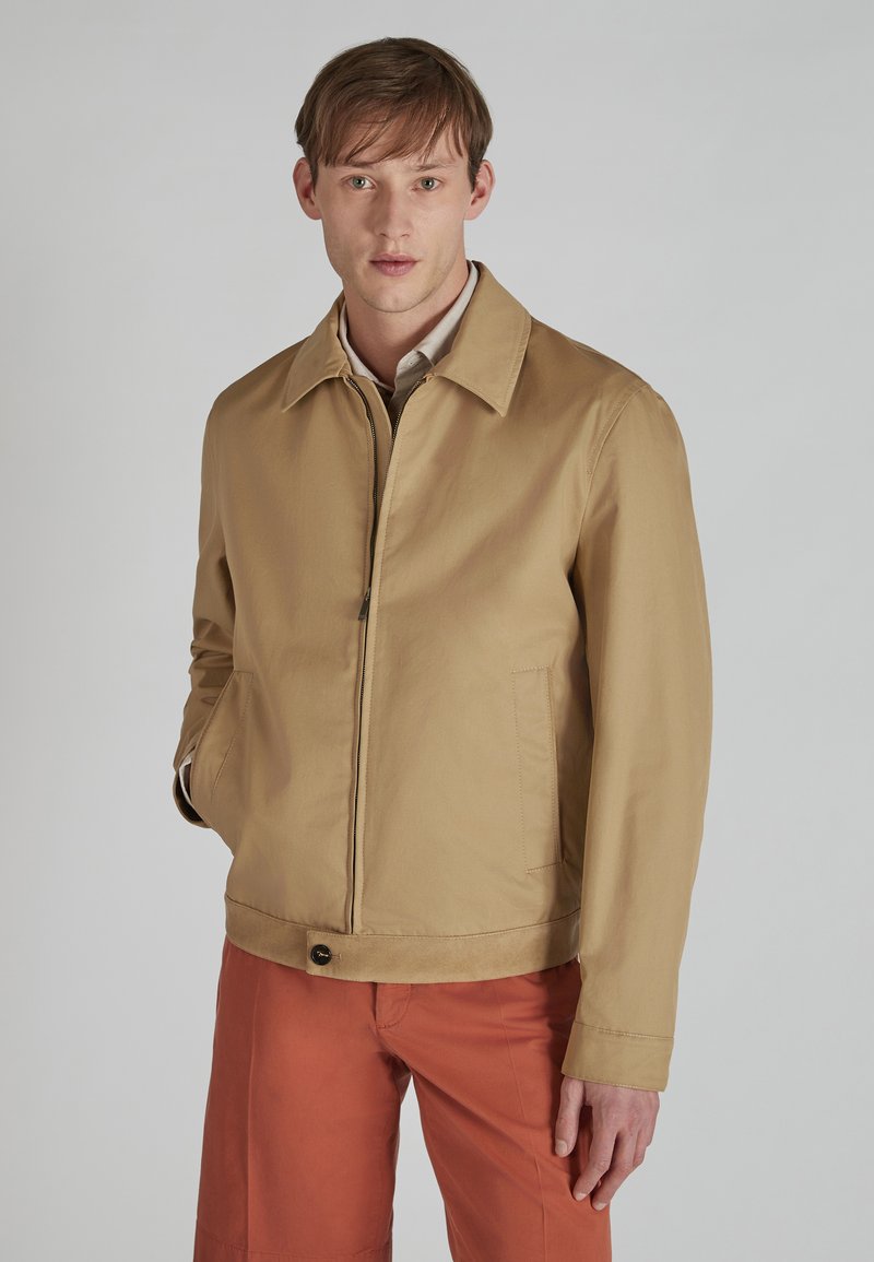 Chaqueta marrón claro con cuello, cremallera frontal y dos bolsillos laterales. Textura suave con un brillo sutil, combinada con pantalones naranjas.