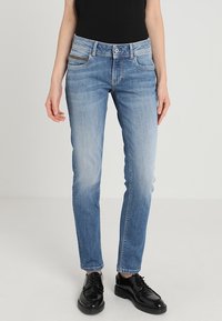 Světle modré džínové kalhoty s vybledlým efektem, úzkým střihem, pěti kapsami a jemnými švovými detaily. Nošeny s černou obuví.