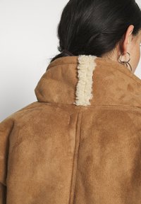 Brun fuskmocka-kappa med hög krage och en strukturerad elfenbensfärgad detalj längs med mitt bak. Slät yta med synliga sömdetaljer.