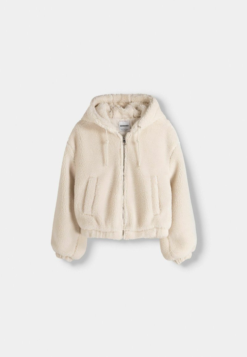Haut à capuche court de couleur crème en tissu pelucheux et texturé. Dispose d'une fermeture éclair avant, de deux poches latérales et d'un capuchon avec cordon de serrage.