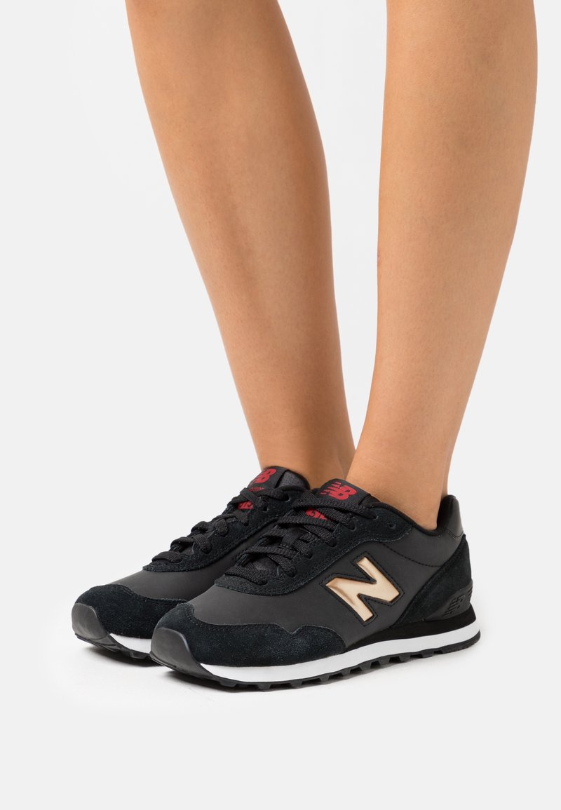 Jambes portant des baskets New Balance noires avec un logo "N" beige et un marquage rouge sur la languette, debout sur un fond blanc.