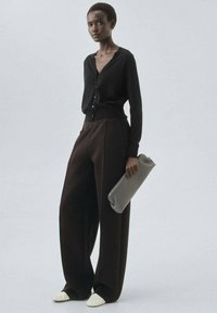 Svart cardigan, vida mörkbruna byxor och skor med krämfärgade toppar. En grå clutch med en jämn textur kompletterar outfiten.