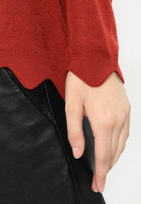 Roter Strickpullover mit gewelltem Saum, der über einem schwarzen Lederrock getragen wird. Nahaufnahme einer Hand, die auf dem Rand des Rocks ruht.