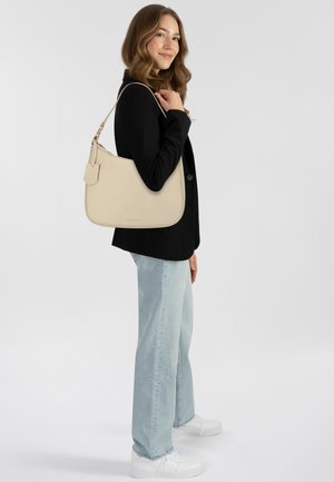 Borsa a tracolla beige in pelle con una texture liscia, forma arrotondata e etichetta decorativa. Indossata sulla spalla, abbinata a jeans azzurri chiari e un blazer nero.