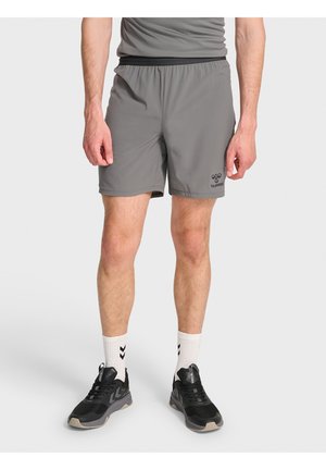 Mężczyzna w szarych sportowych szortach, białych skarpetkach z czarnymi chevronami i czarnych butach do biegania, stojący na jednolitym tle.