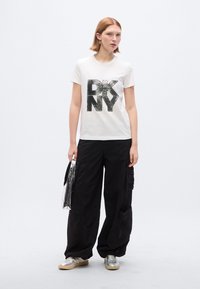 Λευκό βαμβακερό μπλουζάκι με μεγάλο μαύρο γράφημα «DKNY». Συνδυάζεται με φαρδιά μαύρη παντελόνια καρό και ασημένια αθλητικά παπούτσια. Διαφανές τσαντάκι.