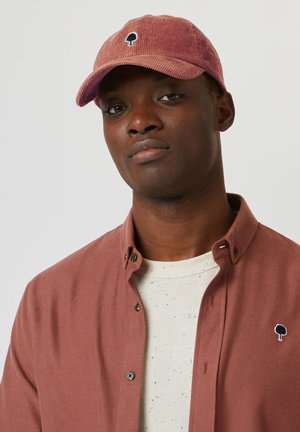 Casquette en velours côtelé bordeaux avec un logo d'arbre brodé, portée avec un t-shirt de couleur claire présentant des specks multicolores et une chemise rose à boutons.