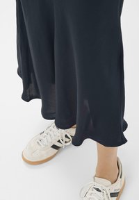 Melna plūstoša kleita ar viļņotu apakšmalu, savienota ar baltām sporta kurpēm ar melniem akcentiem un struktūrizotām zolemi. Gluda auduma drapējums maigi krīt.