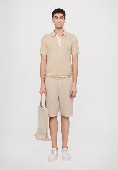 Camisa polo de manga corta beige con cuello, combinada con pantalones cortos de lino beige. La persona sostiene una bolsa de lona beige. Zapatillas blancas completan el conjunto.