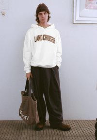 Fehér pulóver "LAND CRUISER" felirattal barna színben, laza fekete nadrággal és sötétbarna csizmával párosítva, barna táskával a kézben.