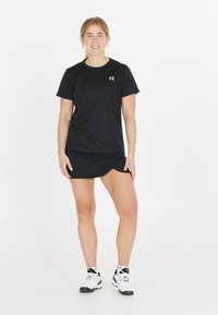 Nuori nainen, joka pukeutunut mustiin urheiluvaatteisiin ja valkoisiin lenkkareihin, seisoo hymyillen yksinkertaista valkoista taustaa vasten.