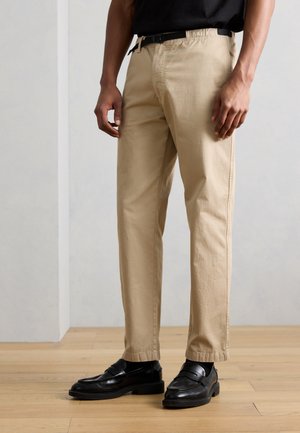 Homme portant un pantalon beige à jambes droites avec une ceinture noire, des mocassins noirs et des chaussettes blanches, debout sur un sol en bois contre un mur gris.